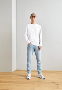 Langärmliges weißes Hemd, hellblaue Jeans und weiße Sneakers mit blauen Akzenten; Modell trägt eine beige Mütze und steht auf einem Holzboden.