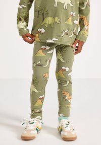 Bambino che indossa una maglietta a maniche lunghe verde con stampa di dinosauri e leggings con motivi di vulcani e palme, scarpe da ginnastica bianche e calzini azzurri chiari.