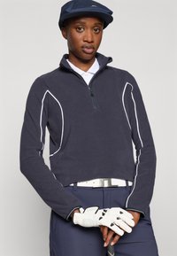 Marineblaue Fleece-Jacke mit weißen Akzenten, langen Ärmeln, hohem Kragen, kombiniert mit einem weißen Poloshirt, marineblauen Hosen und einem weißen Golfhandschuh.