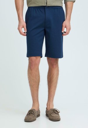 Mann trägt marineblaue knielange Shorts, olivgrünes Hemd zum Hosenbund gesteckt und braune Wildleder-Schnürschuhe, steht vor einem schlichten Hintergrund.