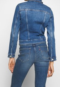 Person wearing a blue denim jacket and matching blue denim jeans, shown from the back against a plain light background.