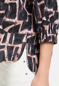 Blusa a maniche lunghe in tessuto leggero con un motivo geometrico in nero, beige e rosso. Presenta polsini elasticizzati e un dettaglio a bottone sul lato.