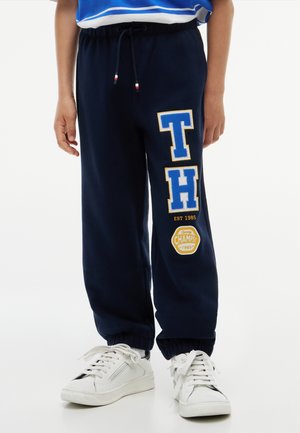 Kind draagt marineblauwe sweatpants met "TH" en "Champs 1985" patches, witte sneakers en een blauw gestreept shirt.