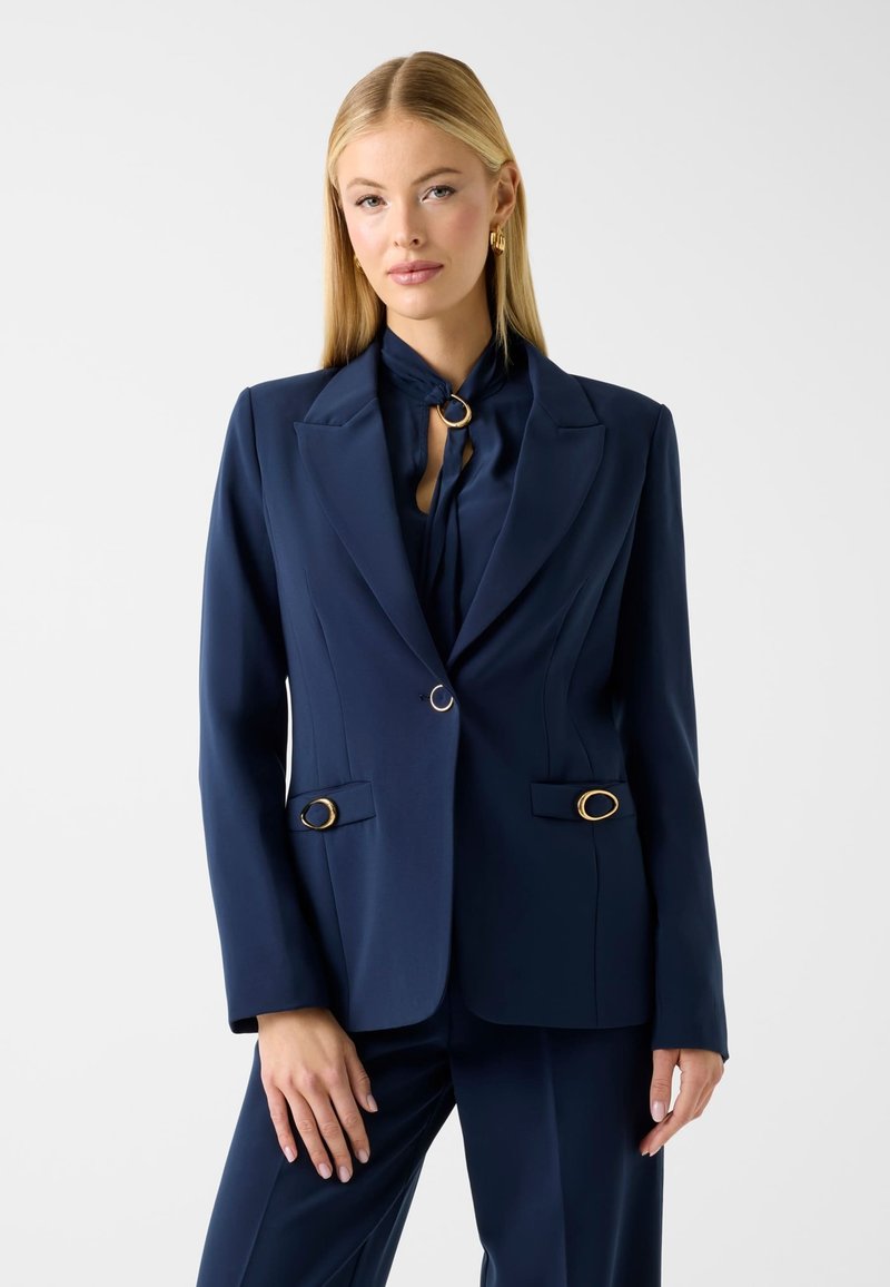 Femme blonde portant un costume sur mesure bleu marine avec des détails en anneaux dorés sur le blazer et un pantalon large assorti, posant devant un fond blanc.