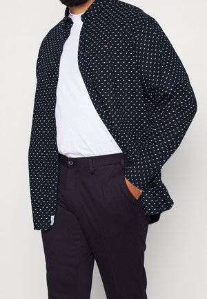 Mann trägt schwarze Hose, weißes T-Shirt und dunkelblaues Hemd mit kleinem weißem Rautenmuster, Hand in der Tasche.