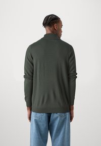 Dunkelgrüner Strickpullover mit hohem Kragen, langen Ärmeln und gerippten Bündchen, kombiniert mit hellblauen Jeans.