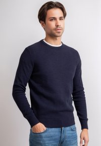 Donkerblauwe gebreide trui met een textuurpatroon, ronde halslijn en ribgebreide manchetten en zoom. Gepaard met lichtblauwe jeans.