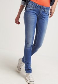 Slim-fit modré džíny v kombinaci s bílými teniskami a oranžovým topem, které ukazují uvolněný módní styl od pasu po nohy.