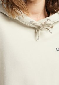 Gros plan d'un sweat à capuche beige avec un cordon noué en noeud près du cou et des cheveux courts châtain clair partiellement visibles au-dessus.