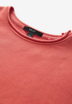 T-shirt color corallo in cotone morbido con scollo a girocollo, dotato di etichetta con il nome del marchio "NEXT" cucita all'altezza del collo.