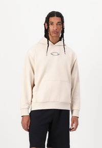 Cremefarvet hætte sweatshirt med kængurulomme og broderet logo på brystet, parret med sorte shorts. Glat, blød tekstur.