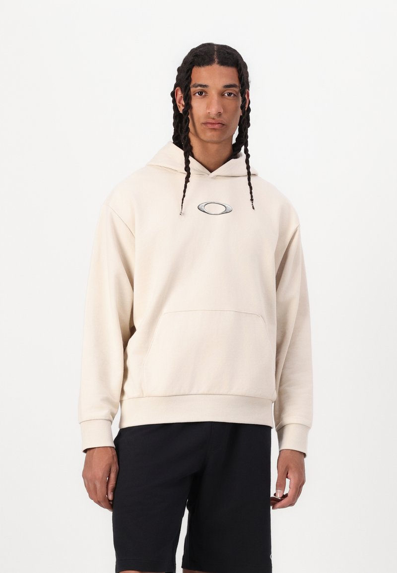 Cremefarvet hætte sweatshirt med kængurulomme og broderet logo på brystet, parret med sorte shorts. Glat, blød tekstur.
