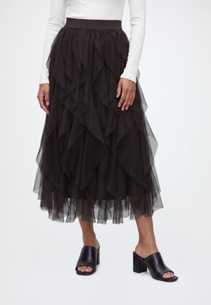 Femme portant une jupe midi en tulle noir à plusieurs couches, un haut blanc à manches longues, et des mules noires à talons ouverts, debout sur un fond blanc.