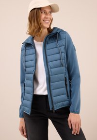 Blauwe gewatteerde jas met een capuchon, rits aan de voorkant en zijzakken. Kenmerkt zich door een gladde buitenstof en elastische manchetten. Draagt over een witte top.