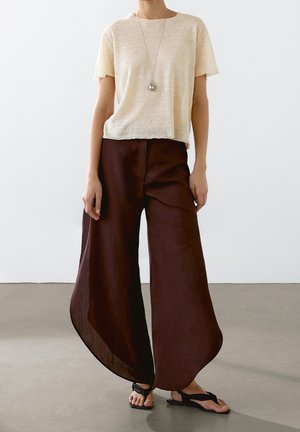 Personne portant un haut beige à manches courtes, un pantalon large marron foncé, des sandales noires et un long collier avec un pendentif rond.