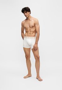 Homme torse nu debout pieds nus portant un boxer blanc avec ceinture dorée à motifs sur fond clair uni.