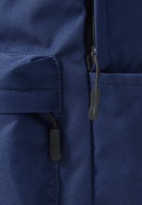 Marineblå ryggsekk i stoff med teksturert overflate, med svart glidelås og dragere, samt en sidelomme for oppbevaring.