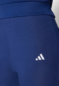Sötétkék sportleggingszek, testhezálló szabással, sima felülettel és fehér Adidas logóval az alsó oldalon.