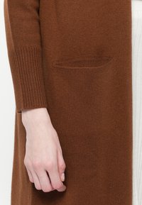 Cardigan lavorato a maglia marrone con polsini a coste e una tasca frontale. Texture morbida, maniche lunghe e design aperto, indossato sopra un capo più leggero.
