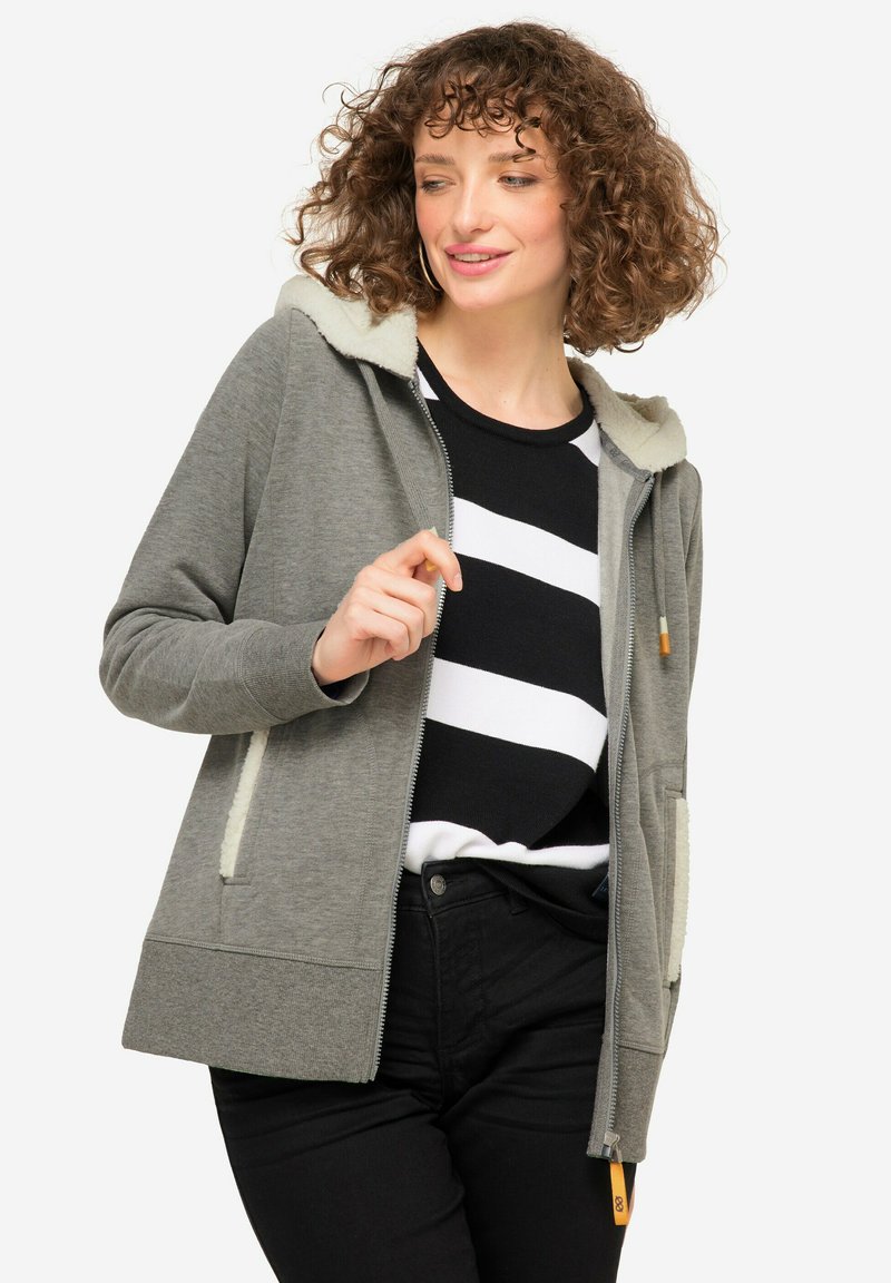 Felpa grigia con zip e cappuccio foderato in pile crema, con tasche laterali, indossata sopra una maglia a strisce bianco e nero, che mette in evidenza un look informale.