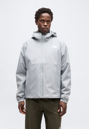 Jeune homme portant une veste de pluie grise à capuche et un pantalon vert olive, debout devant un fond clair uni.