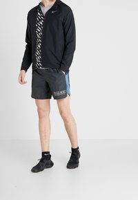 Zwarte lichtgewicht jas met een patroon aan de voorkant, gecombineerd met donkere shorts met blauwe accenten en witte branding. Zwarte sportschoenen.