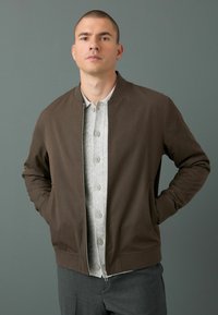 Chaqueta bomber marrón con cremallera frontal, bolsillos laterales y una camisa de botón gris debajo. Textura de tejido simple y suave con puños acanalados.
