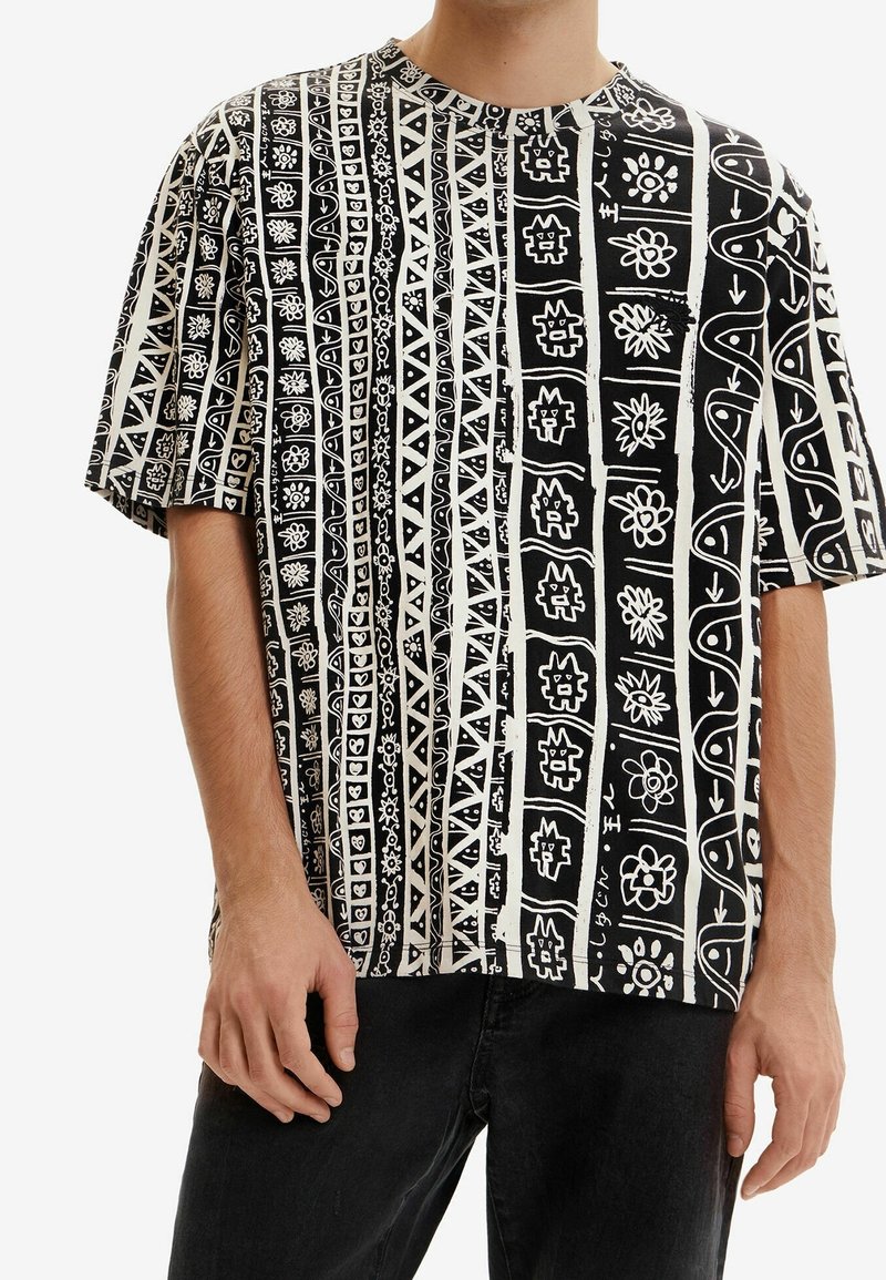 T-shirt noir et blanc à manches courtes avec des motifs tribaux et floraux verticaux porté par une personne en pantalon noir.