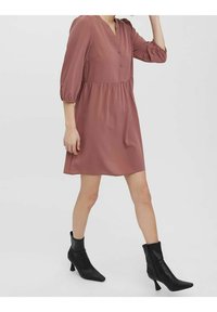 Rosekleurige jurk met een V-hals, knoopdetails en driekwart pofmouwen. Gepaard met zwarte enkellaarzen met een spitse neus.