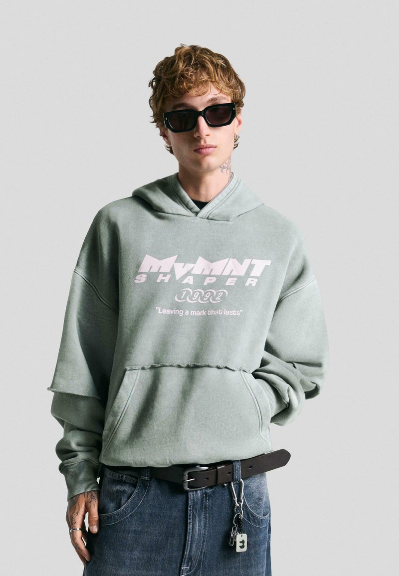 Vihreä mintunvärinen oversized huppari, jossa on graafinen teksti, risaiset helmat ja kiristysnauha hupussa. Yhdistetty tummiin farkkuihin ja mustaan vyöhön, jossa on metalliosia.