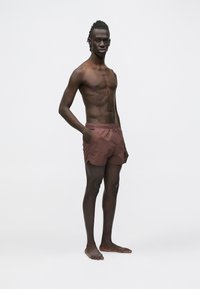 Brązowe kąpielówki z elastycznym paskiem w talii i bocznymi kieszeniami, prezentowane na stojącym męskim modelu z gołym torsorem na tle jednolitego tła.