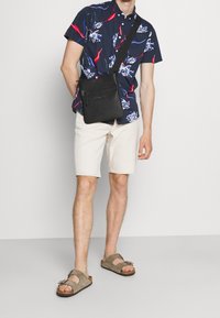 Bolso cruzado sintético negro con bolsillo frontal con cremallera, llevado sobre una camisa de manga corta floral navy y pantalones cortos beige, combinado con sandalias marrones.