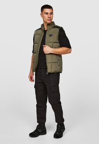 Olijfgroene puffer gilet met een rits aan de voorkant en een staande kraag, gedragen over een zwart T-shirt. Gecombineerd met zwarte cargobroeken en sneakers.
