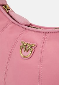 Różowa torba nylonowa z emblematem w kształcie podwójnego ptaka w złotym odcieniu oraz okrągłym detalem w kształcie pierścienia. Widoczny zamek błyskawiczny i szyte akcenty. Gładka tekstura.