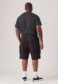 Svarte cargo-shorts med sidelommer, kombinert med en grå t-skjorte. Hvite sokker og svarte sneakers fullfører antrekket. Enkel bakgrunn.