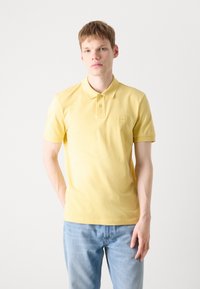 Gul polo-skjorte med klassisk krage, korte ermer, en liten logo på brystet, sammen med lyseblå denimjeans.