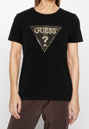Person iført en sort kortærmet skjorte med et gyldent paliet-trekantet Guess-logo og mørkebrune bukser.