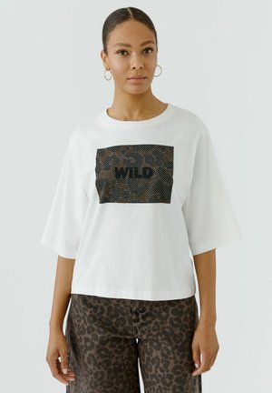 Vrouw draagt een wit los t-shirt met een zwart en bruin luipaardprint rechthoek en het woord "WILD,", gecombineerd met broeken met luipaardprint.