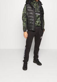 Sort polstret vest over en grøn camo-mønstret top, parret med sorte sweatpants med hvide grafiske detaljer og sorte sneakers.