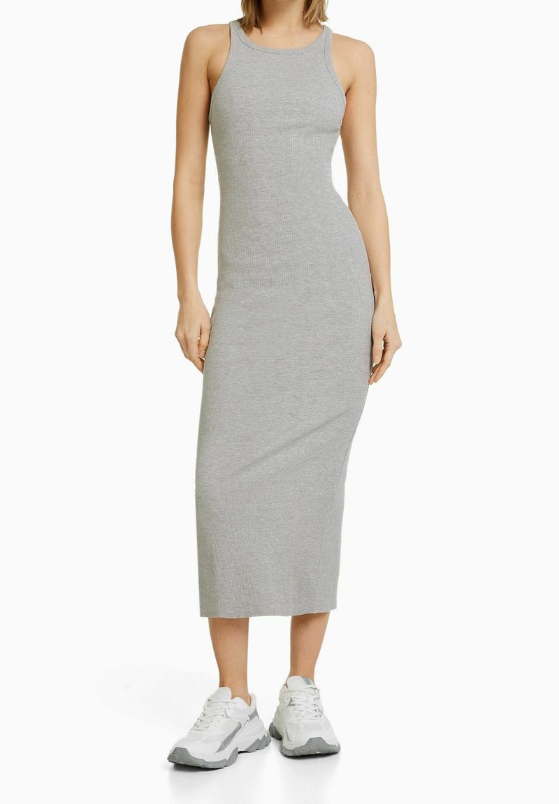 Femme portant une robe midi grise cintrée sans manches associée à des baskets blanches sur fond blanc.