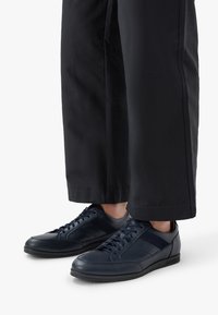 Námořnické kožené boty s texturovaným svrškem, kulatou špičkou a plochou černou podrážkou. V kombinaci s širokými černými kalhotami, z nichž je vidět pouze dolní část.