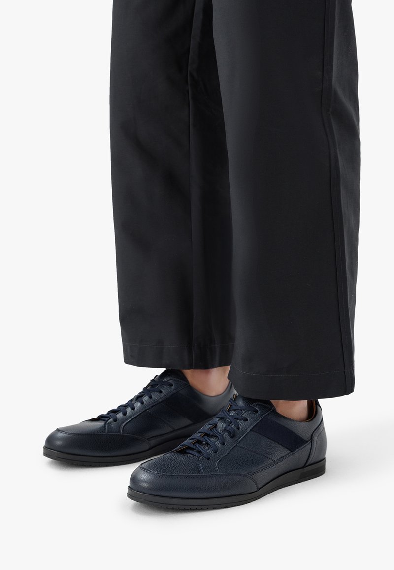 Námořnické kožené boty s texturovaným svrškem, kulatou špičkou a plochou černou podrážkou. V kombinaci s širokými černými kalhotami, z nichž je vidět pouze dolní část.