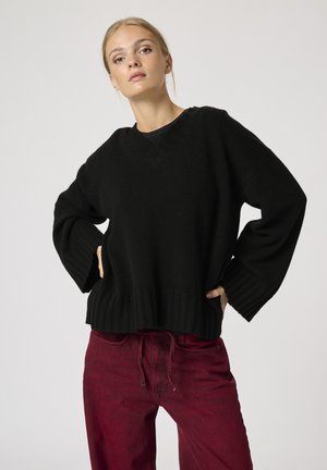 Fekete, oversized pulóver V-nyakkal, bordázott mandzsettákkal és szegéllyel. Magas derekú piros farmernadrággal párosítva. Simább textúra és kényelmes illeszkedés.
