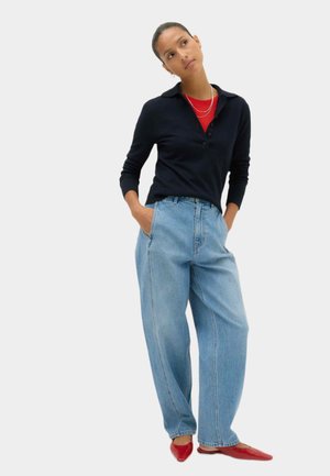 Vrouw staat met handen in de zakken van lichtblauwe wijdvallende jeans, draagt een zwarte top met lange mouwen, een rood overhemd, rode schoenen en zilveren kettingen.