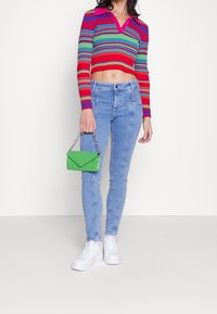 Červený a vícebarevný pruhovaný krátký svetr s výstřihem do V, kombinovaný s světle modrými přiléhavými džínami. V ruce drží malou zelenou kabelku s řetízkovým uchem.