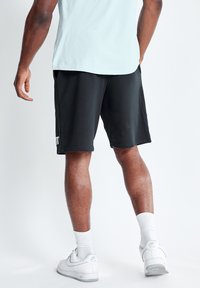 Pantalones cortos deportivos negros hechos de una tela suave. Presentan un corte holgado, bolsillos laterales y un logo blanco en la pierna izquierda. Se llevan con zapatillas blancas.