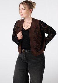 Blonde vrouw die gouden creolen, gelaagde kettingen, een zwart kanttop, een bruin geborduurd vest, een zwarte riem en donkere high-waisted jeans draagt, poseert.