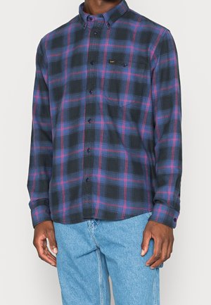 Chemise à manches longues boutonnée avec un motif à carreaux bleu et rouge, portée non rentrée dans un jean bleu clair sur une personne debout.
