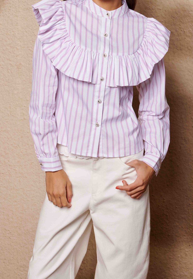 Femme portant un chemisier rayé verticalement rose et blanc avec un empiècement volanté et un pantalon blanc taille haute, debout contre un mur beige.