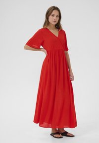 Robe maxi rouge avec un décolleté en V et des manches courtes, présentant une taille cintrée et un tissu texturé. Associée à des sandales noires simples.